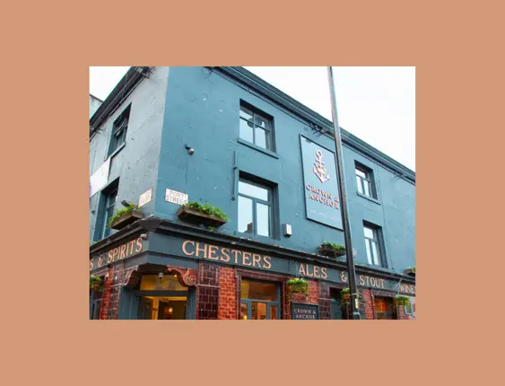 A corner pub painted blue - with a sign saying "Crown &amp; Anchor" on the facing side. It has a banner wrapping around it, listing Ales, Stout, Wines &amp; Spirits.