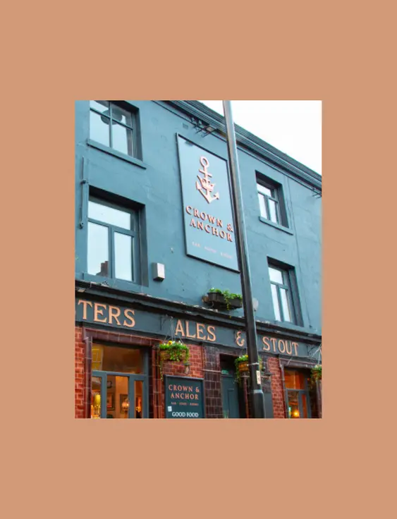 A corner pub painted blue - with a sign saying "Crown & Anchor" on the facing side. It has a banner wrapping around it, listing Ales, Stout, Wines & Spirits.